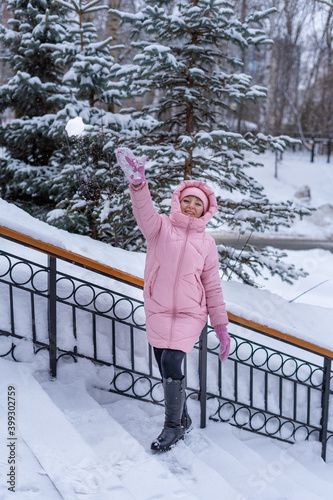 Young woman in a pink down jacket in a city park standing on the stairs throws snowballs