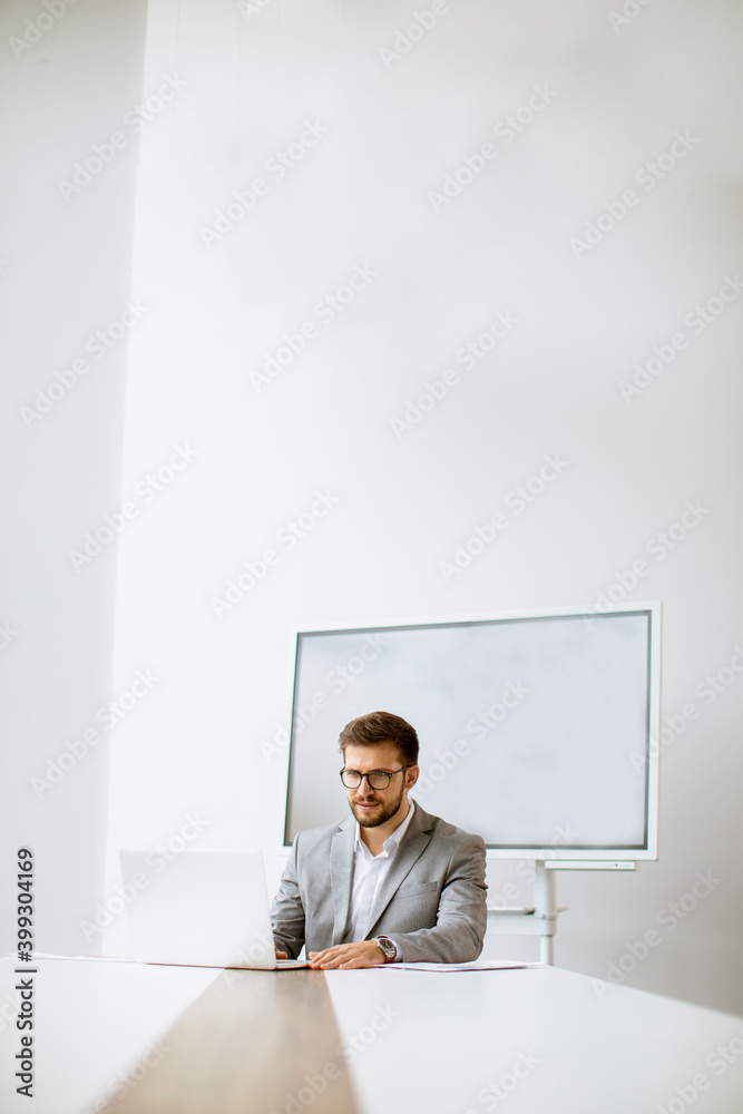 Young man working on laptop in bright office with big screen behind him Young man working on laptop in bright office with big screen behind him