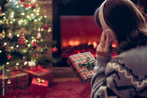 Woman in headphones with christmas gift box in hand sitting on fluffy plaid near fireplace and christmas tree.