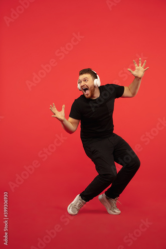Dancing expressive. Full length portrait of young high jumping man gesturing isolated on red studio background. Attractive male caucasian model. Copyspace. Human emotions, facial expression concept. © master1305