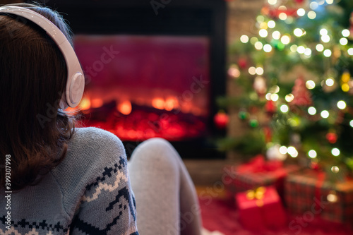 Alone sad woman in headphones sitting and warming at winter evening near fireplace flame and christmas tree.