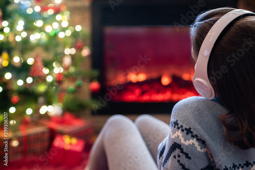 Woman in headphones sitting and warming at winter evening near fireplace flame and christmas tree.