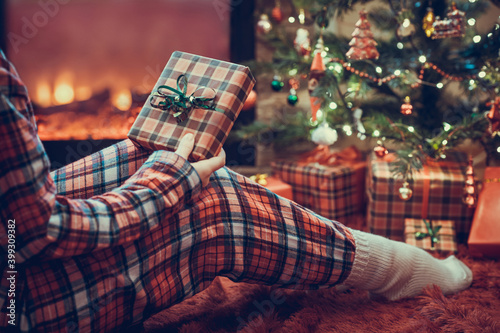 Woman in pajamas with christmas gift box in hand sitting on fluffy plaid near fireplace and christmas tree.