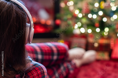 Woman in pajama in headphones lying and warming at winter evening near fireplace flame and christmas tree.