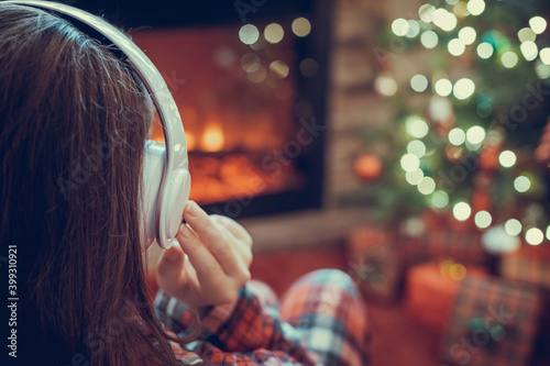 Woman in pajama in headphones sitting and warming at winter evening near fireplace flame and christmas tree.