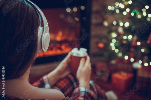 Woman in pajama in headphones with cup of hot cocoa and marshmallow sitting and warming at winter evening near fireplace flame and christmas tree.