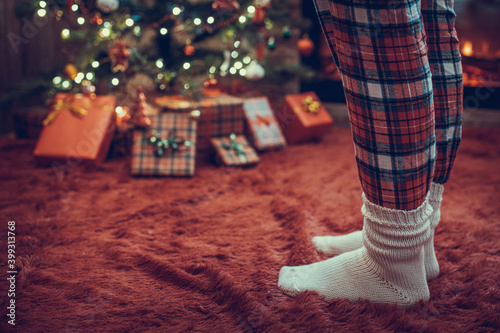 Female feet in pajama and winter socks standing near a Christmas tree with gifts and fireplace at morning. Concept