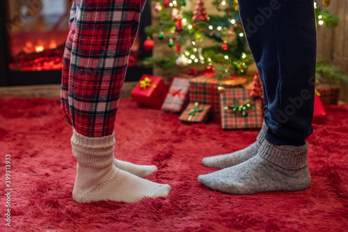 Woman feet standing in tip toe in winter socks on male lags on a fluffy red blanket near a Christmas tree with gifts. Concept