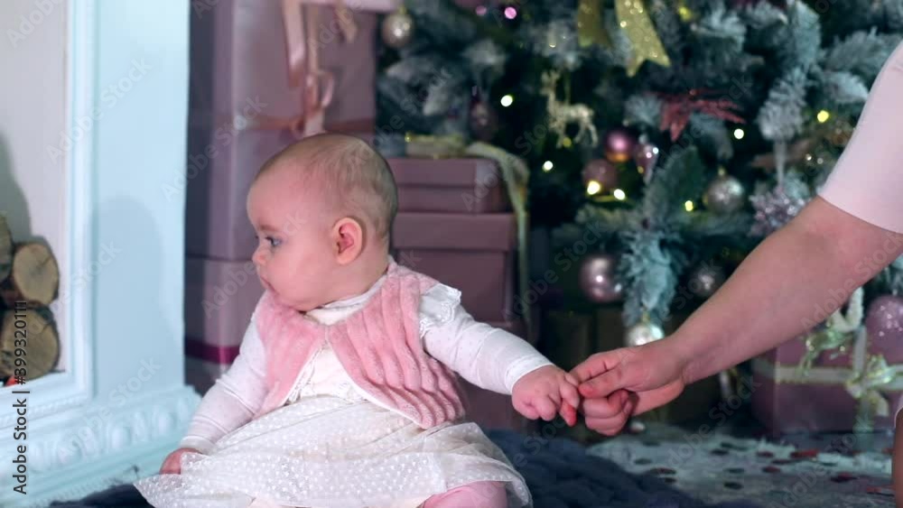 Cute little stylish baby girl in white dress sitting on carpet near Christmas tree enjoying New Year celebration at home with family. Caucasian female child. Home modern interior. Lights on background Cute little stylish baby girl in white dress sitting on carpet near Christmas tree enjoying New Year celebration at home with family. Caucasian female child. Home modern interior. Lights on background