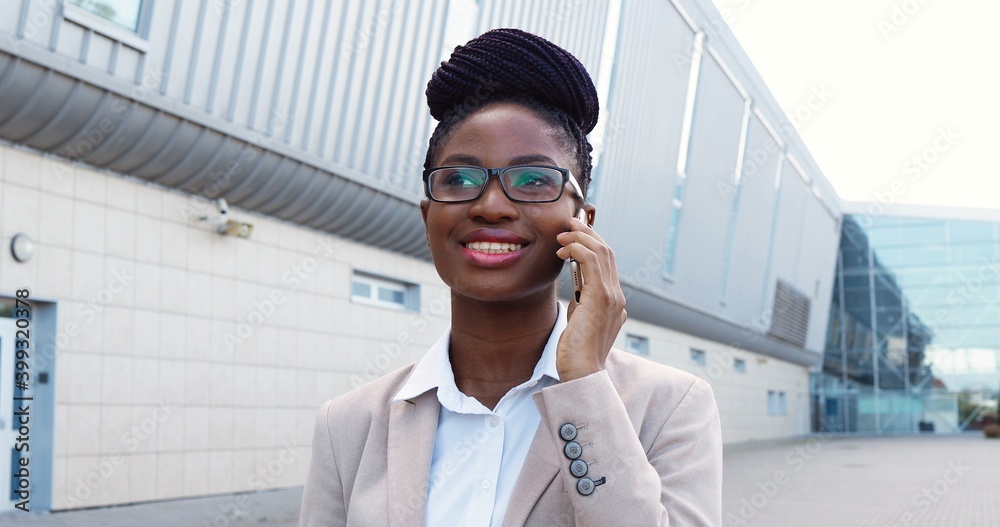 African American young happy businesswoman in suit and glasses talking on cellphone and standing outdoors. Good-looking woman in business style speaking on mobile phone. African American young happy businesswoman in suit and glasses talking on cellphone and standing outdoors. Good-looking woman in business style speaking on mobile phone.