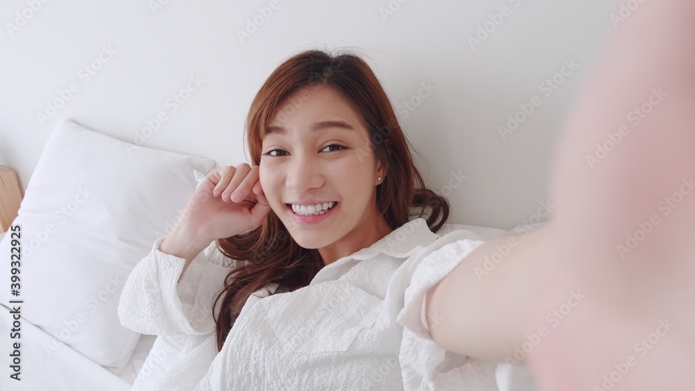 ฺBeautiful Asian girl smiling and show her hand in a white shirt takes a video selfie with a friend on the bed in a white room. ฺBeautiful Asian girl smiling and show her hand in a white shirt takes a video selfie with a friend on the bed in a white room.