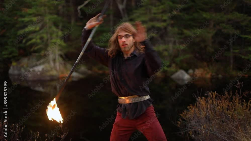 Handsome young man with long hair holding and spinning fire stick during fire jamming standing near lake side in a forest in Canada Handsome young man with long hair holding and spinning fire stick during fire jamming standing near lake side in a forest in Canada