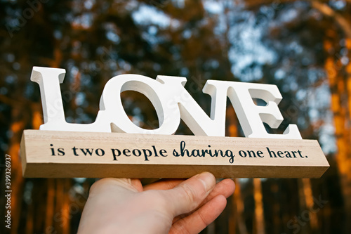 a sign made of wood with the inscription about the hearts of lovers, held by a man's hand, against the background of the forest