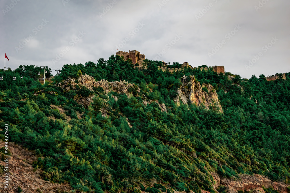 Alanya Castle (Turkey) on the top of a mountain among a green forest - bottom view. Ancient fortress with a tower and stone walls among green trees on a hill in a summer-autumn cloudy day Alanya Castle (Turkey) on the top of a mountain among a green forest - bottom view. Ancient fortress with a tower and stone walls among green trees on a hill in a summer-autumn cloudy day