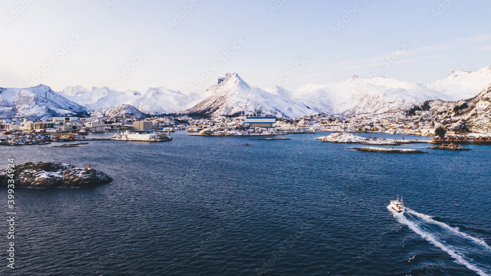 Aerial view of ferry boat crossing cold nordic water surface to fishing village in wild environment near breathtaking fjords, Scandinavian environment with snowy mountains on horizon Aerial view of ferry boat crossing cold nordic water surface to fishing village in wild environment near breathtaking fjords, Scandinavian environment with snowy mountains on horizon