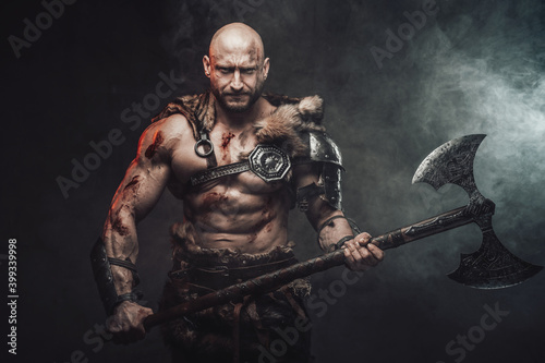 Atmospheric portrait of a powerful and hairless scandinavian warrior posing in dark and smokey background holding his steel two handed axe looking at camera.