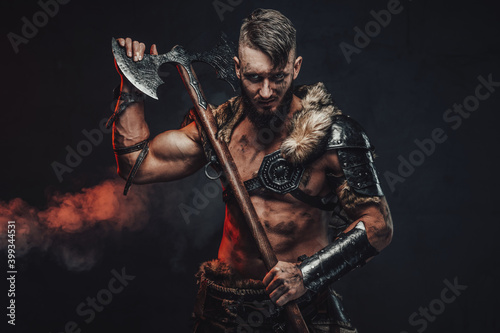 Dressed in light armour with fur scandinavian seafarer armed with two handed axe poses in dark background.