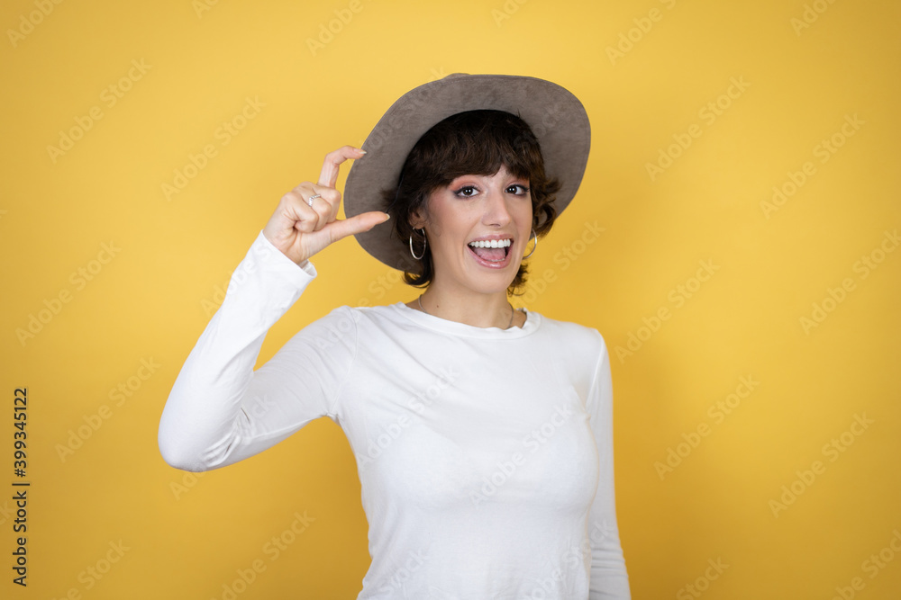 Young caucasian woman wearing hat over isolated yellow background smiling and confident gesturing with hand doing small size sign with fingers . Measure concept. Young caucasian woman wearing hat over isolated yellow background smiling and confident gesturing with hand doing small size sign with fingers . Measure concept.
