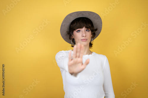 Young caucasian woman wearing hat over isolated yellow background serious and doing stop sing with palm of the hand.