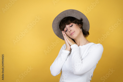 Young caucasian woman wearing hat over isolated yellow background sleeping tired dreaming and posing with hands together while smiling with closed eyes.