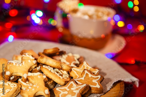 Christmas gingerbread cookies on a wooden stand and a cup of hot chocolate with marshmallows in the back, on a red background with a beautiful bokeh of glowing garland. Christmas and New Year concept.