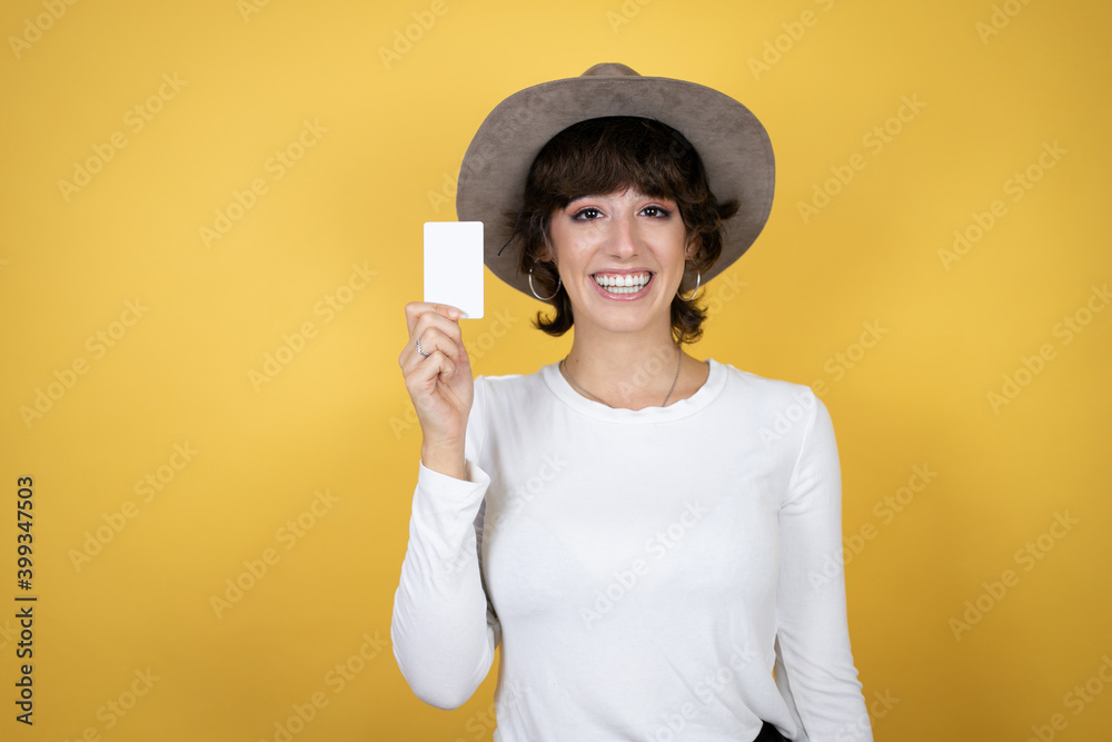 Young caucasian woman wearing hat over isolated yellow background smiling and holding white card Young caucasian woman wearing hat over isolated yellow background smiling and holding white card