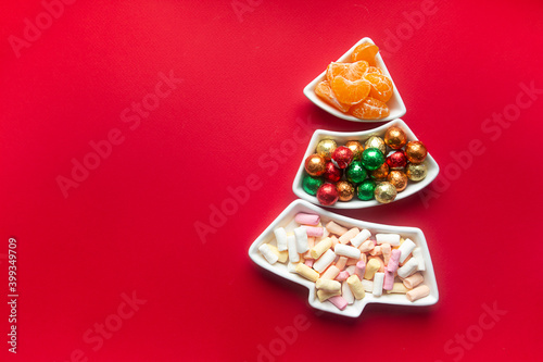 on a red background there is a tray in the form of a Christmas tree. sweets in the tray: marshmallows, round candies in foil and tangerine slices