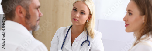 Charming young woman in white lab coat looking at camera and holding pen while sitting at the table with coworkers photo