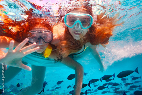 Two beautiful girls in scuba mask swim underwater and wave hand to the camera smiling in the pool