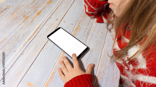 The child is sitting at a table with a phone with a white screen. A girl with a phone layout. In new year's clothing. New Year