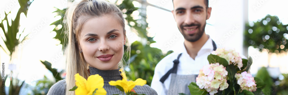 Charming young woman and bearded man in apron looking at camera and smiling while demonstrating flowers in plant store Charming young woman and bearded man in apron looking at camera and smiling while demonstrating flowers in plant store