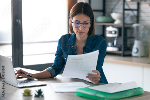 Pretty young business woman working with computer while consulting some invoices and documents in the kitchen at home.