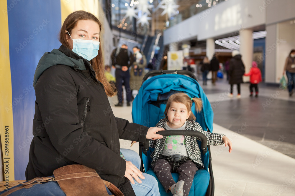 Caucasian mother resting with her daughter sitting in children carriage, family walking in shopping mall, adult woman wearing face mask