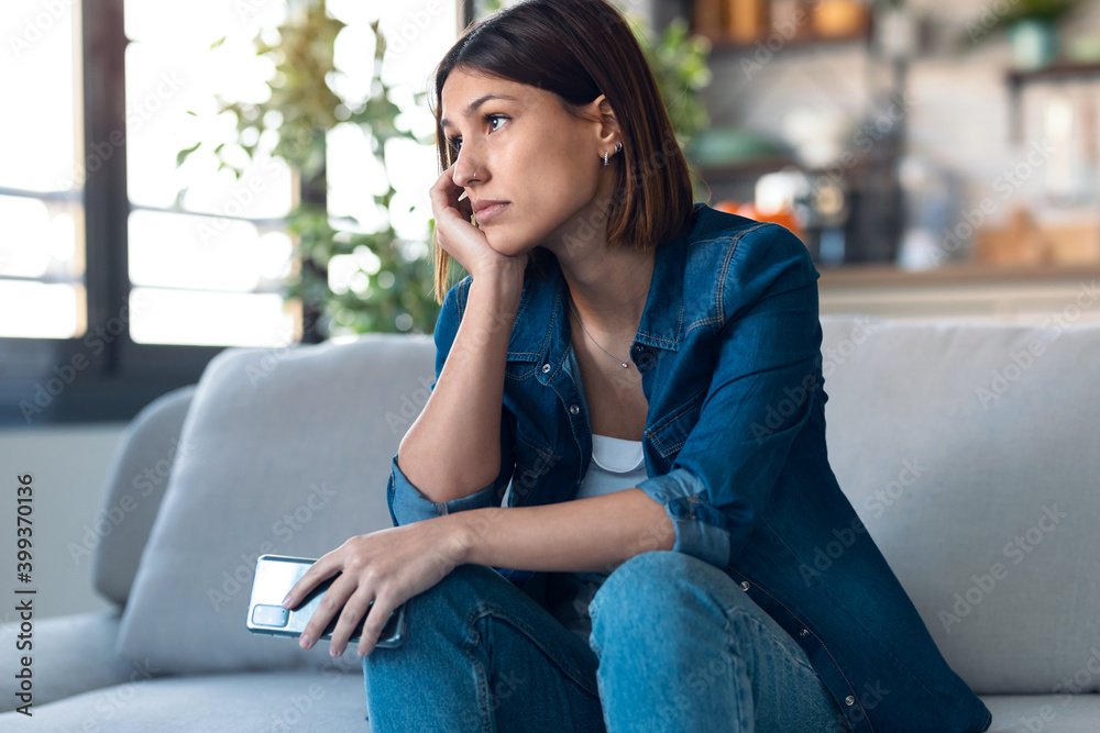 Worried young woman using her mobile phone while thinking about problems sitting on couch in the living room at home. Worried young woman using her mobile phone while thinking about problems sitting on couch in the living room at home.