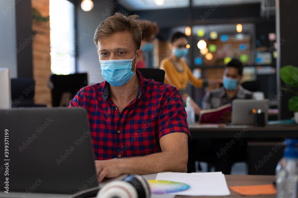 Caucasian man wearing face mask using laptop at desk in casual office