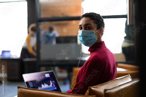 Portrait of mixed race woman wearing face mask working on laptop wearing earphones in casual office
