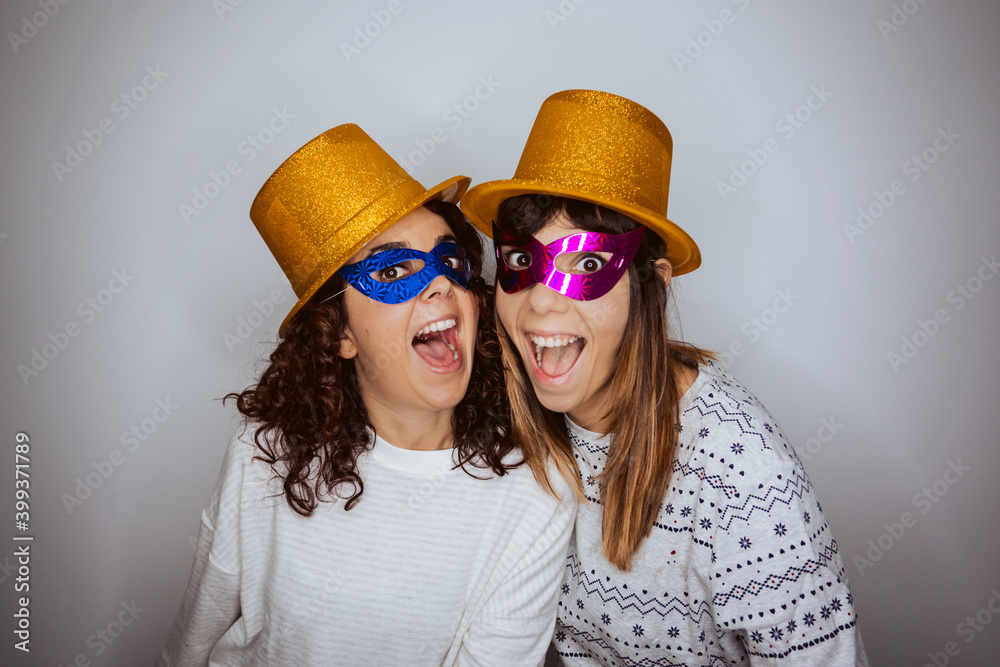 .Two beautiful women saying goodbye to the year 2020 in pajamas from home due to the pandemic caused by covid19. Festive and fun attitude wearing golden hat and colorful mask .Two beautiful women saying goodbye to the year 2020 in pajamas from home due to the pandemic caused by covid19. Festive and fun attitude wearing golden hat and colorful mask