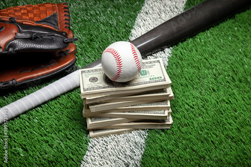 Baseball on top of stack of money with mitt and bat on green field with stripe