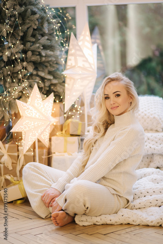 Blond hair woman in white knitted suit sitting at home by Christmas tree. Beautiful woman at winter holidays, Christmas mood
