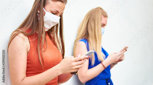Young woman friends with face masks using social media networks with mobile smart phones - Millennial girls keeping distance to prevent the spread of virus - New normal lifestyle concept