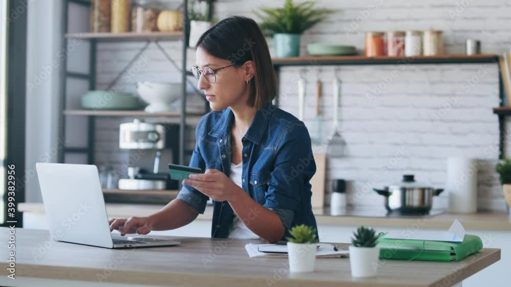 Video of cute young woman holding white credit card for shopping online with computer while sitting in the kitchen at home.
