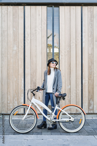 Portrait young pretty Caucasian girl, dressed in a gray jacket, a white sweater and a hat with a bike ride in the city