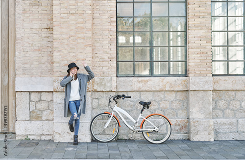 Portrait young pretty Caucasian girl, dressed in a gray jacket, a white sweater and a hat, leaning against the wall with a bike for a ride in the city, talking on the mobile phone