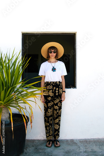 beautiful girl standing near window and palm in ethno clothes and with big hat