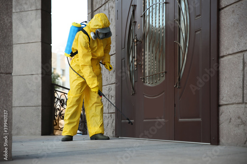 Person in hazmat suit disinfecting entrance door with sprayer. Surface treatment during coronavirus pandemic