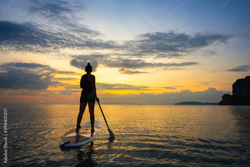 Silhouette of Confidence Asian woman standing on sup board with paddleboarding passing through beautiful sea and twilight sky at summer sunset. Female enjoy recreation water sport in holiday vacation