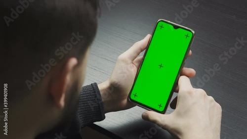 Man looking at screen of mobile phone - shot over the shoulder. Smartphone vertical orientation with a green screen.