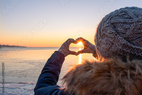 A girl in winter clothes makes a heart with her hands in gloves. Against the background of the frozen sea at sunset. Sun rays. A knitted hat. Love for nature