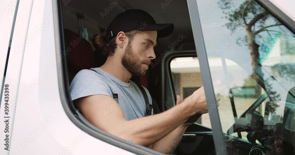 Side view. Close up of Caucasian young handsome male courier sitting on front seat in van tapping and browsing online on tablet device. Delivering orders. Delivery man in uniform typing on gadget Side view. Close up of Caucasian young handsome male courier sitting on front seat in van tapping and browsing online on tablet device. Delivering orders. Delivery man in uniform typing on gadget