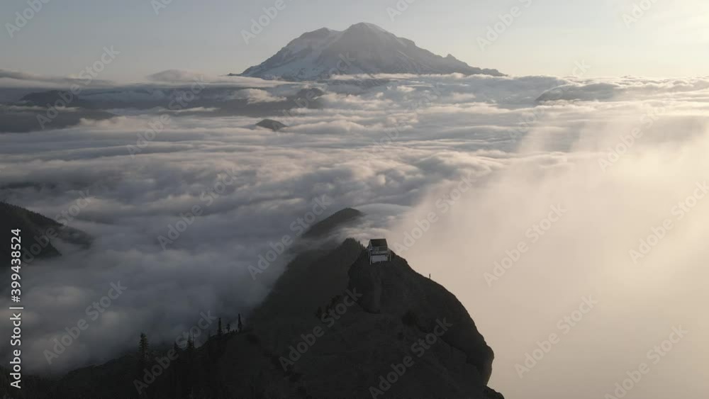 Aerial shot of tiny house standing on top off hill over the cloudscape and beautiful Rock Mountain in background.Sunset in Washington State,United States America. Aerial shot of tiny house standing on top off hill over the cloudscape and beautiful Rock Mountain in background.Sunset in Washington State,United States America.